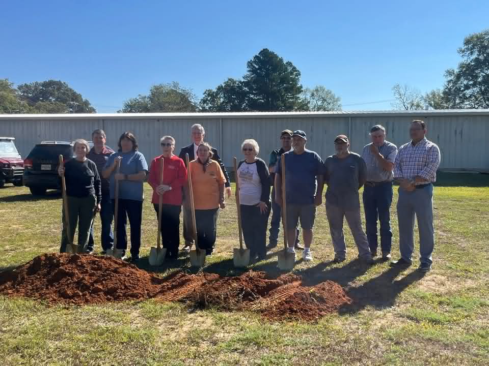 Falkner Heritage Museum Project board members, volunteers and Town of Falkner officials standing behind a line of turned red earth with shovels, at the groundbreaking for the Community Center.